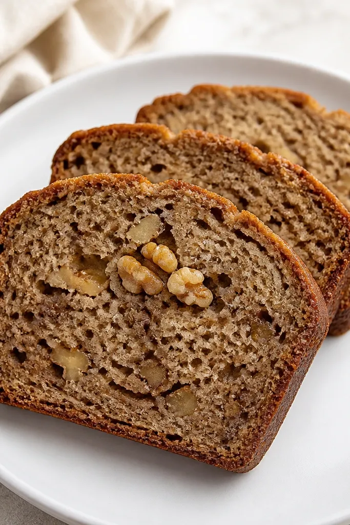 Three slices of banana bread on a white plate, with visible walnut chunks in the crumb; the slices are arranged slightly overlapping, and a few walnut pieces are centered in the front slice.