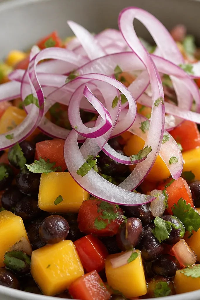 Close-up of salsa with thin, curled red onion ribbons on top. The ribbons look translucent at the edges, and chopped cilantro is scattered like green confetti over mango and black beans.