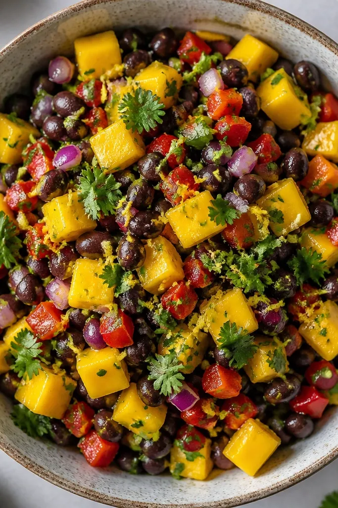 Top-down view where lime zest looks like tiny green-yellow threads across the salsa. Mango cubes are shiny, black beans are visible, and cilantro sits in scattered dots.