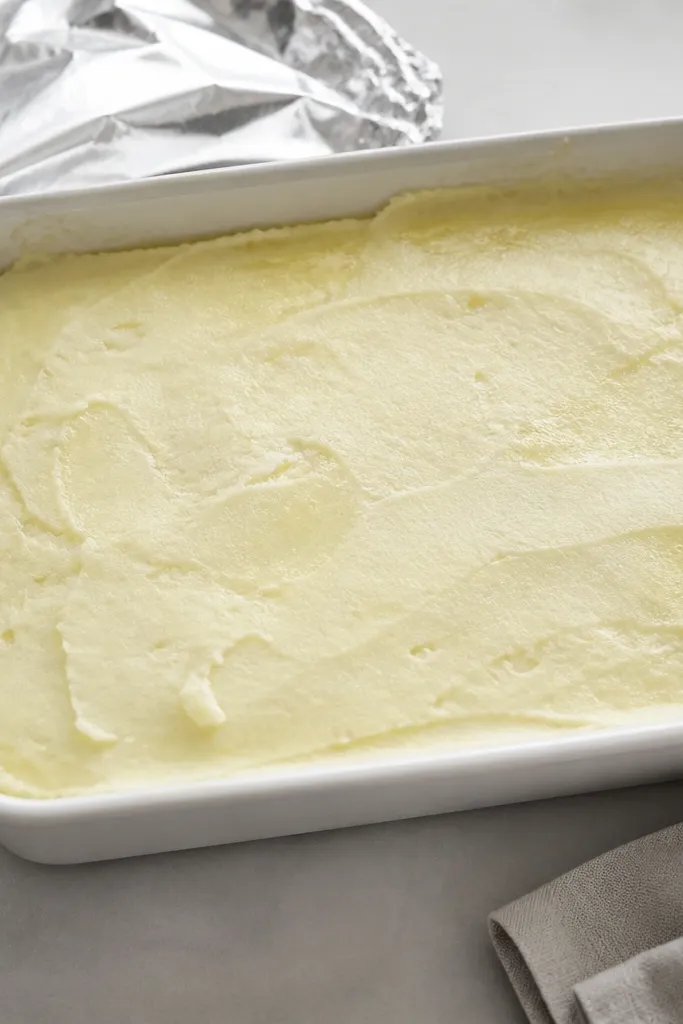 A baking dish filled with smooth mashed potatoes, covered with foil. The top is flat and creamy, with a faint sheen from butter, and edges show a gentle, even thickness.