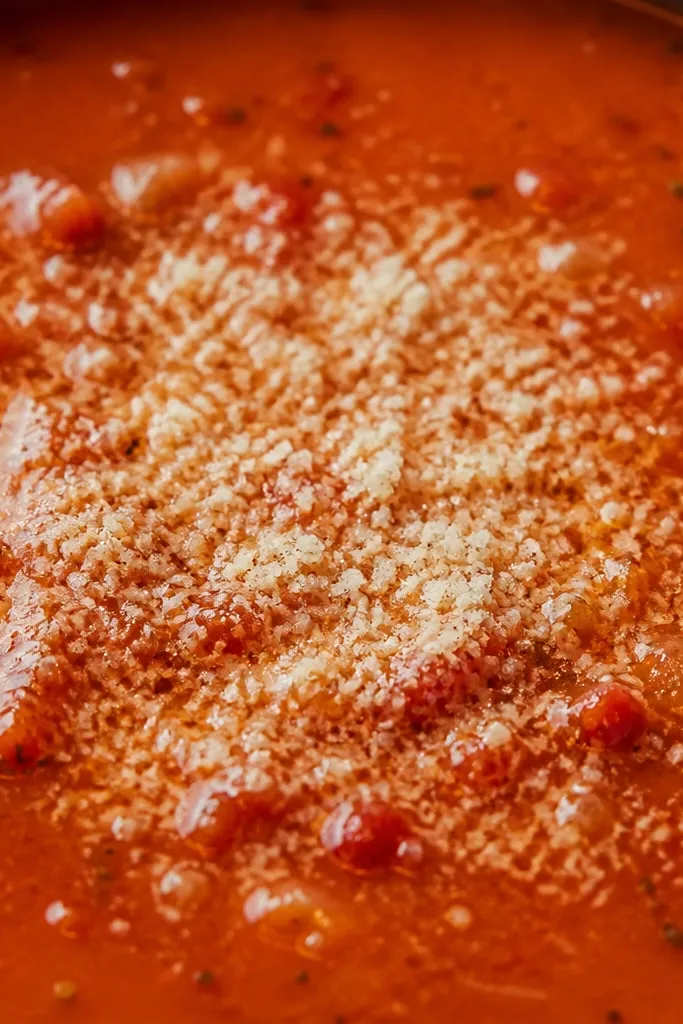 Close-up of soup surface with fine grated parmesan dusting the red broth. The parmesan forms tiny pale specks and melts slightly at the edges.