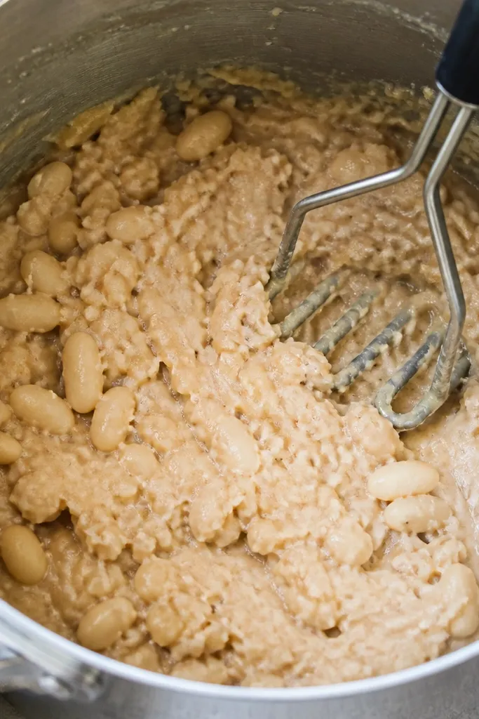 A close-up of a pot where a portion of beans is visibly mashed. You can see a few whole beans still intact, while the rest looks creamy and broken. A potato masher sits against the side of the pot.