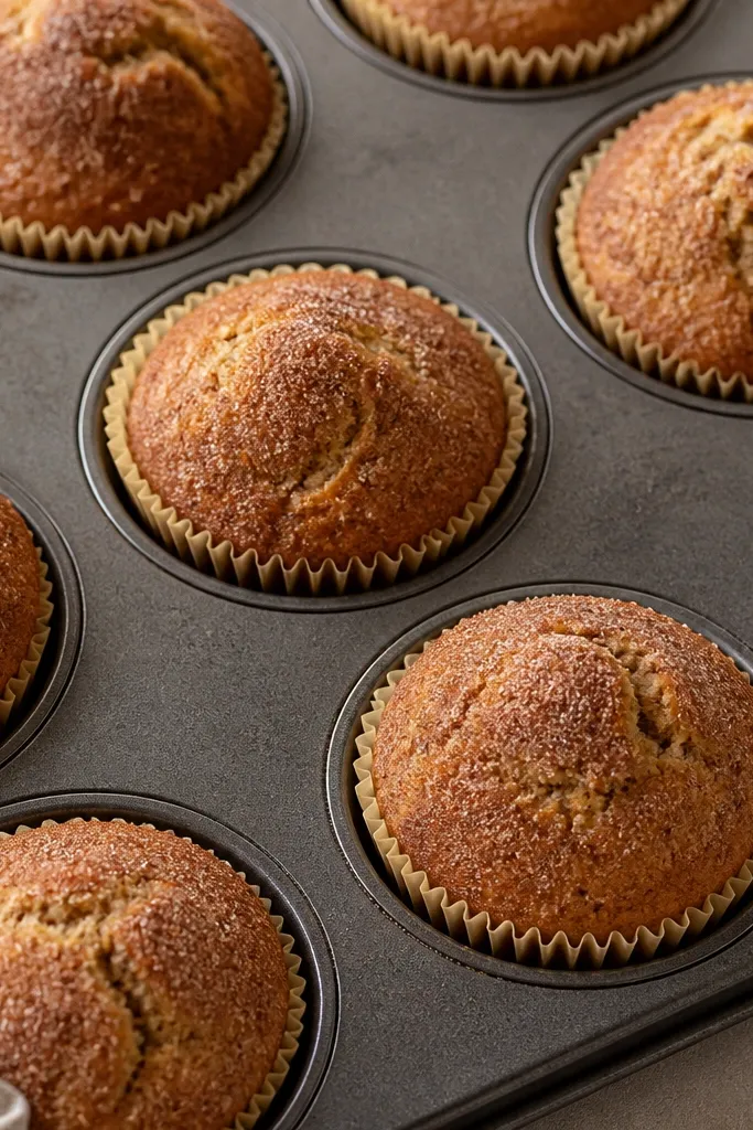A muffin tray lined with light beige paper liners. The muffins have domed tops dusted with cinnamon-brown sugar, with a few small cracks where sugar melted and set.