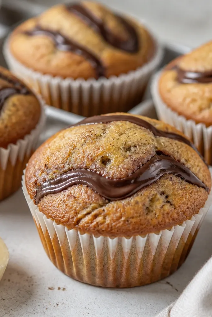 Muffins with a thin chocolate ribbon swirl running across the top. The crumb looks speckled with banana, and the chocolate looks glossy and slightly set.