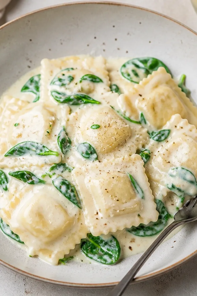 Creamy alfredo ravioli with small green spinach leaves folded through the sauce. The spinach stays bright and doesn't look dark or slimy.