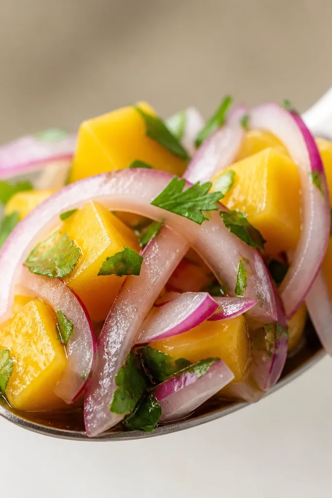 Close-up of salsa on a spoon showing translucent red onion strands and cubes of mango. The liquid around the spoon is minimal, with a light lime sheen instead of a pool.
