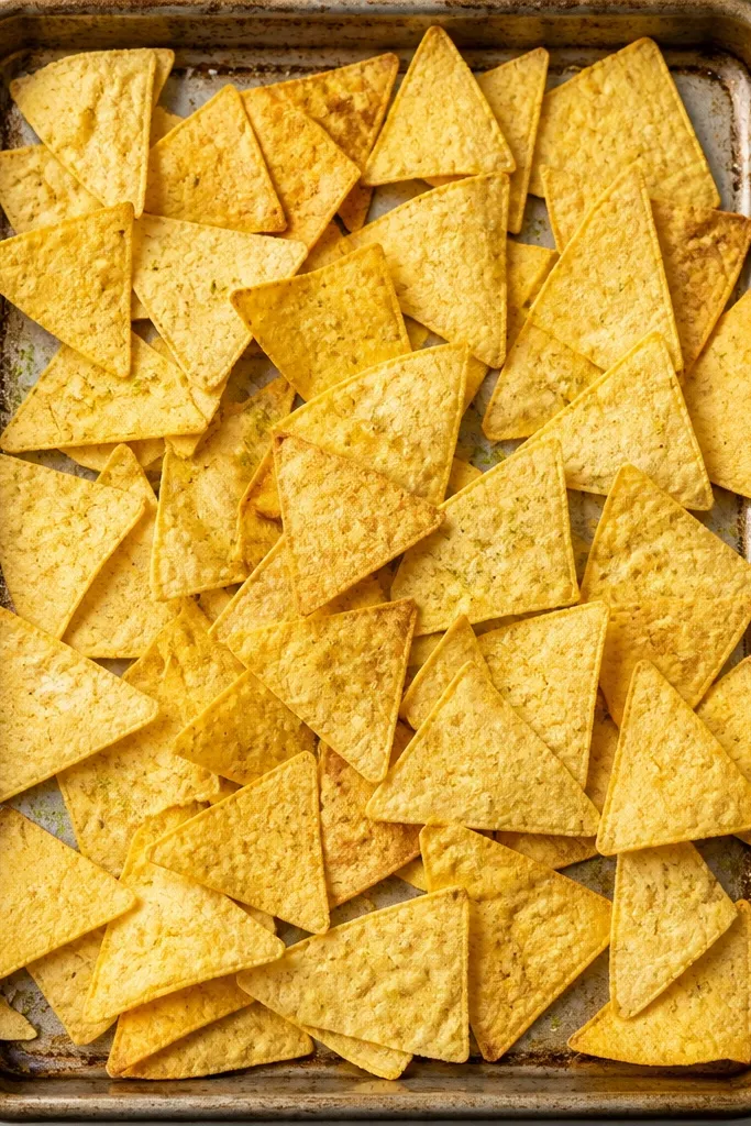 A metal sheet pan covered in a single, even layer of thick tortilla chips, with visible tiny pale-green lime-salt flecks sprinkled across the top.