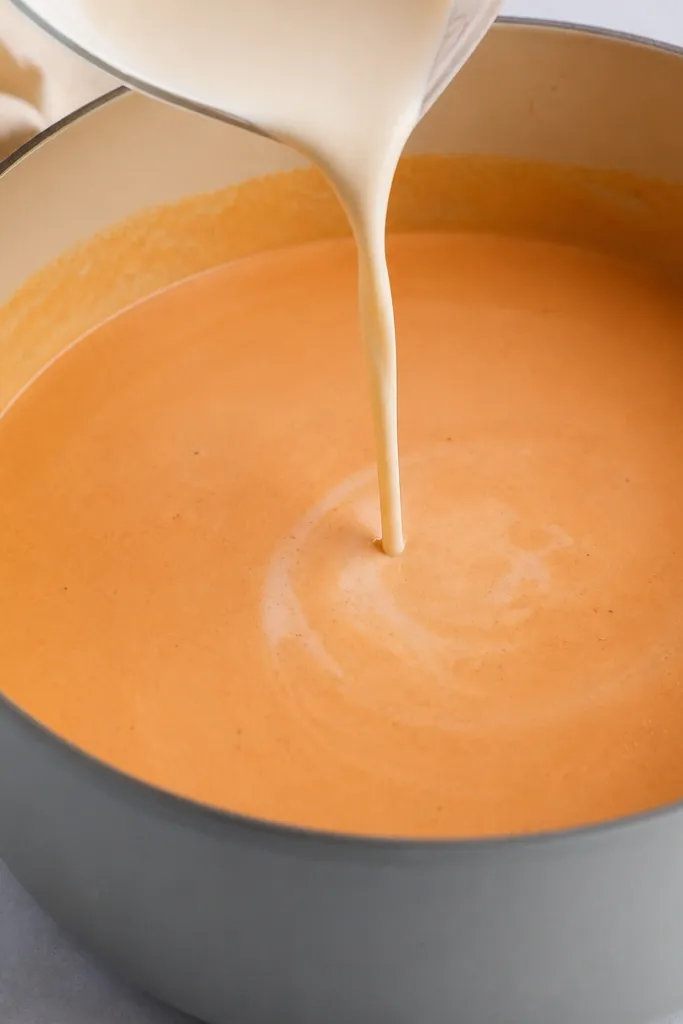A pot with the heat turned off. The cream is poured in a thin stream into the reduced vodka-tomato base. The mixture turns lighter and looks smooth, with no visible curds.