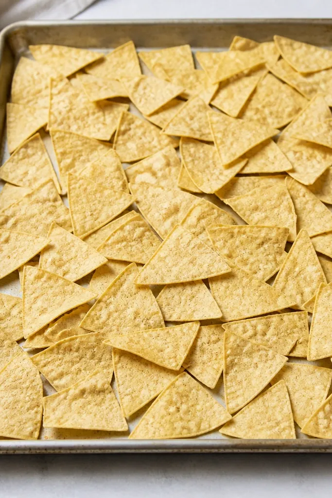 A large sheet pan filled with tortilla chips spread in a mostly even layer, with visible chip edges creating a light golden grid across the pan. No toppings are on yet; the chips look dry and flat.