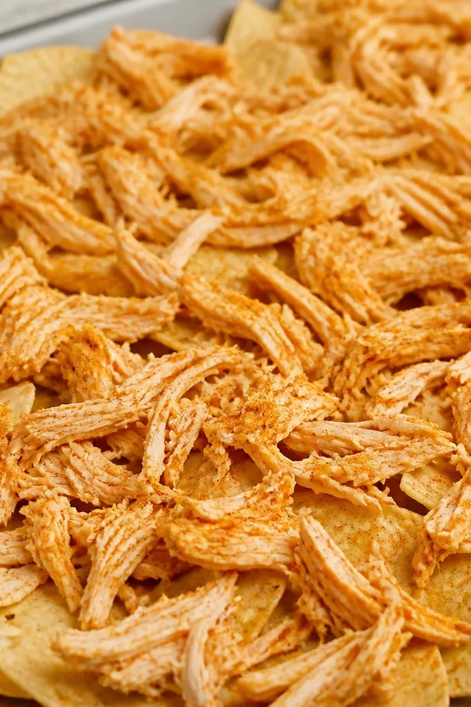 A close-up of shredded chicken pieces spread thin over chips, with small 1/2-inch strands visible and evenly distributed. The chicken looks seasoned with a light orange-red dusting.