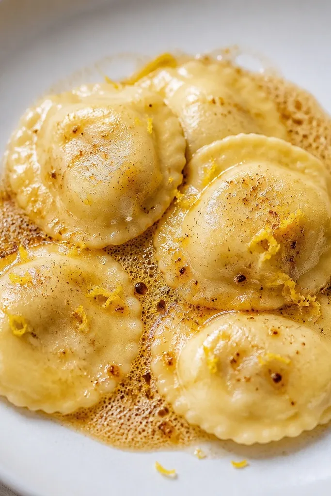 Ravioli plated on a white dish with a thin sheen of brown butter and scattered lemon zest. The butter looks golden and slightly foamy around the edges.
