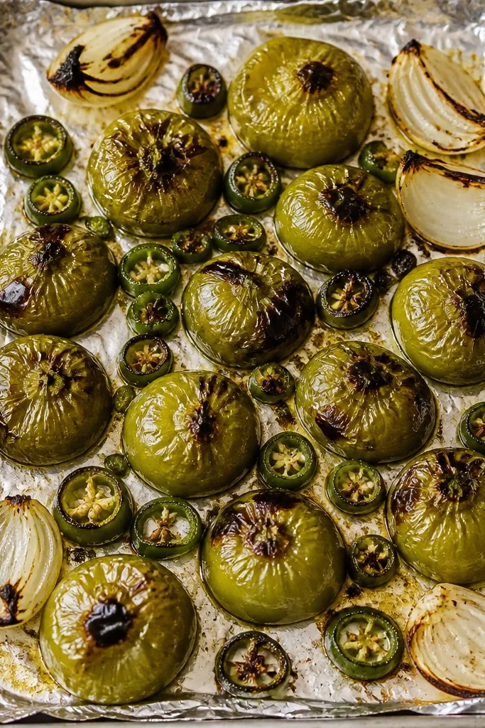 A foil-lined sheet pan with 12 blistering tomatillos, charred green jalapeño rounds, and a few onion wedges around the edges; everything looks glossy and browned with dark freckles.